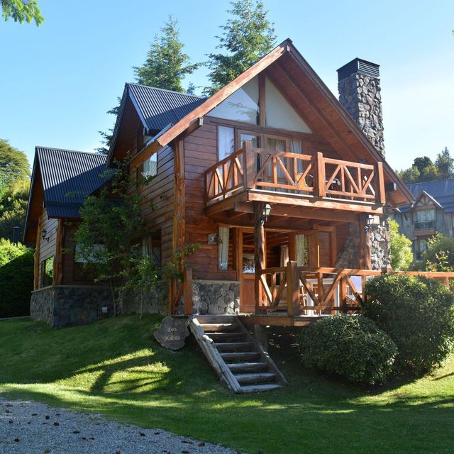 Casa de madera estilo chalet, con balcón, chimenea de piedra y escalones de entrada en un jardín verde y soleado