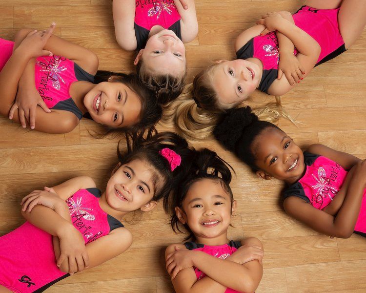 Girls in pink leotards smile, lying in a circle on a wooden floor.
