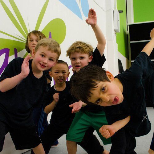 Group of children posing playfully in a dance studio, wearing black tops and various bottoms, colorful wall.