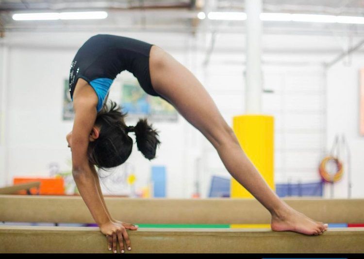 Gymnast arched backwards on balance beam. Blue leotard, indoor gym setting.