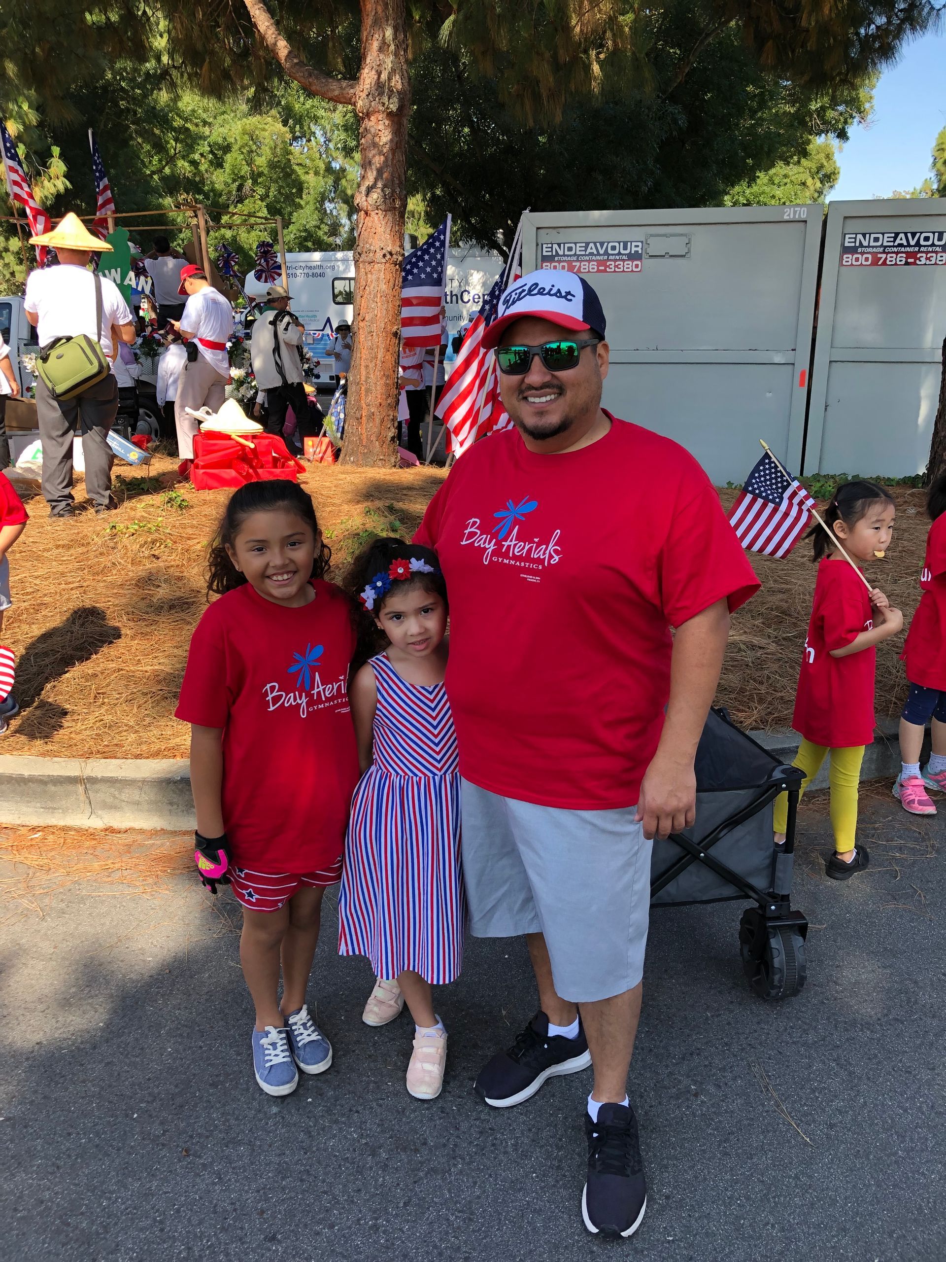 Man and two young girls in red shirts, posing with a stroller, American flags in background.