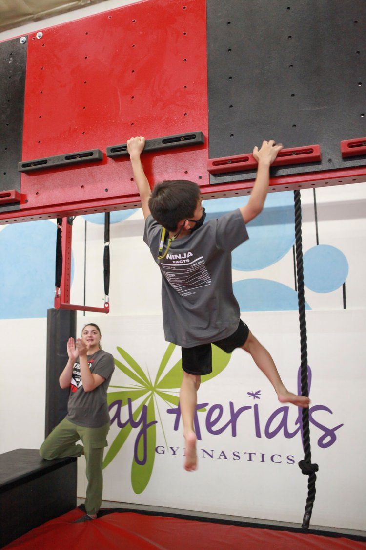 Boy swinging from a climbing wall; red, black, and white gym setting; person watching.