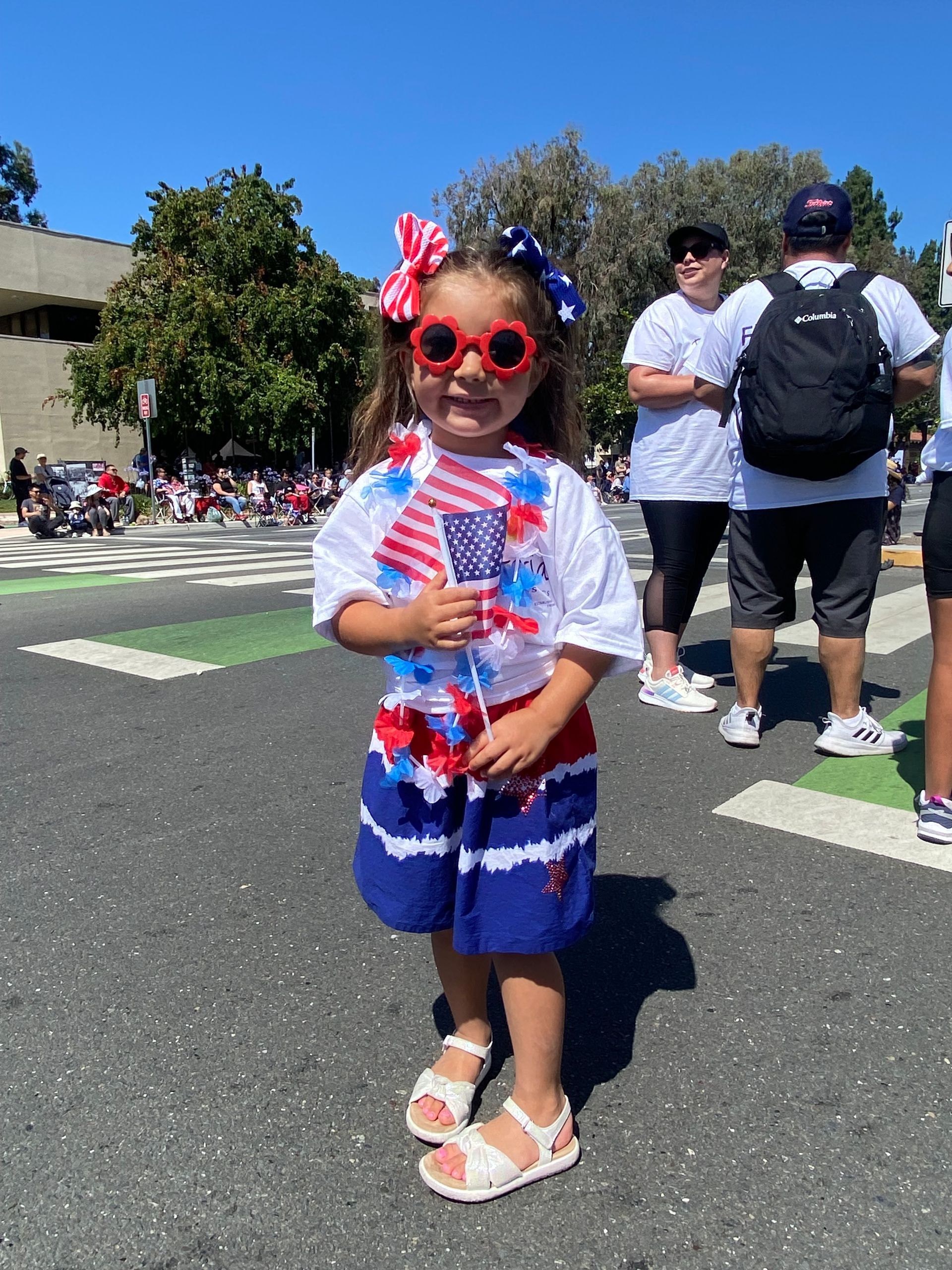 Young child in patriotic attire, holding a small flag, at an outdoor parade.