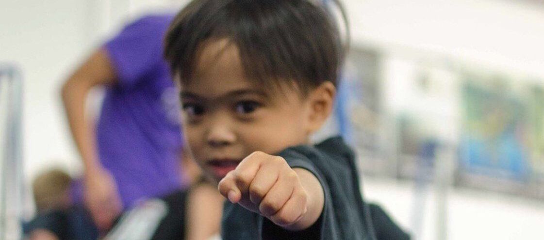 Young child in a black outfit, fist clenched, focused gaze; blurred background of a gym.