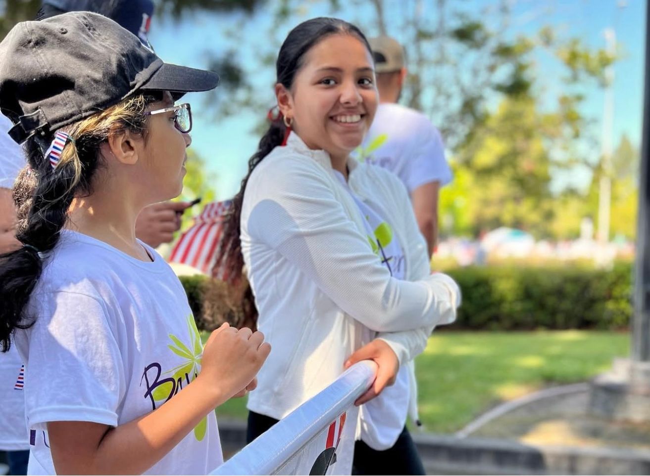 Two young people smiling outdoors, one in a white shirt and the other in a black cap.