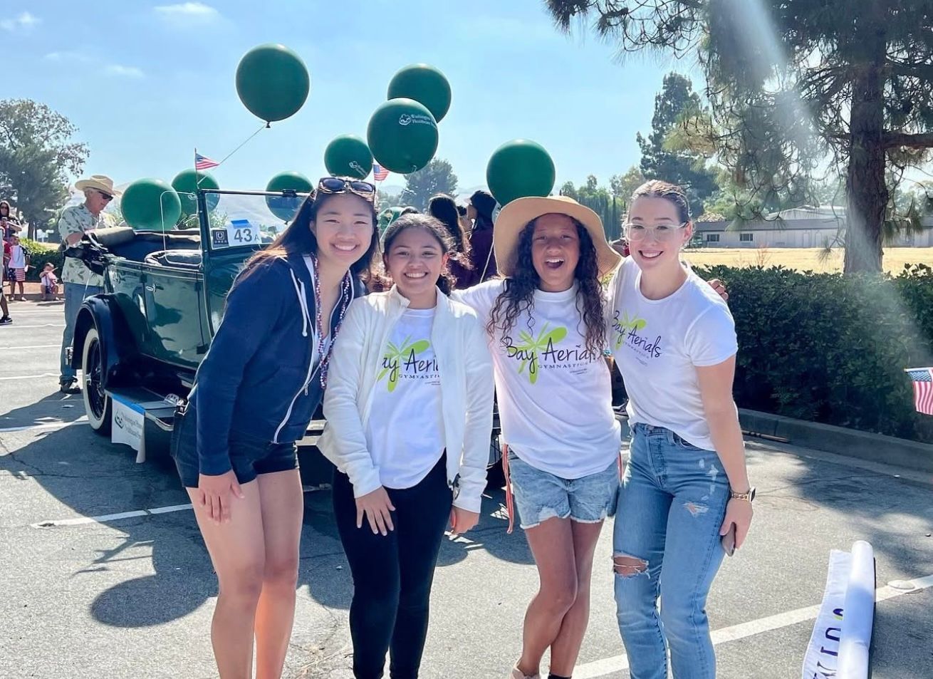 Four people pose in front of a vintage car with green balloons. They wear matching t-shirts, smiling outside.