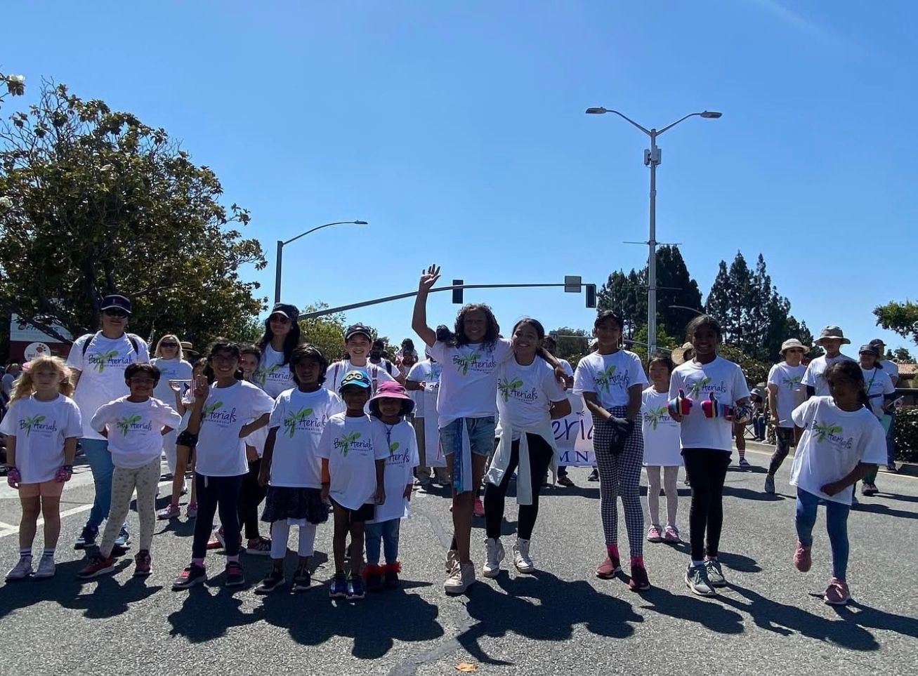 Children and adults in white shirts with a logo walking on a street, waving with smiles. Sunny day.
