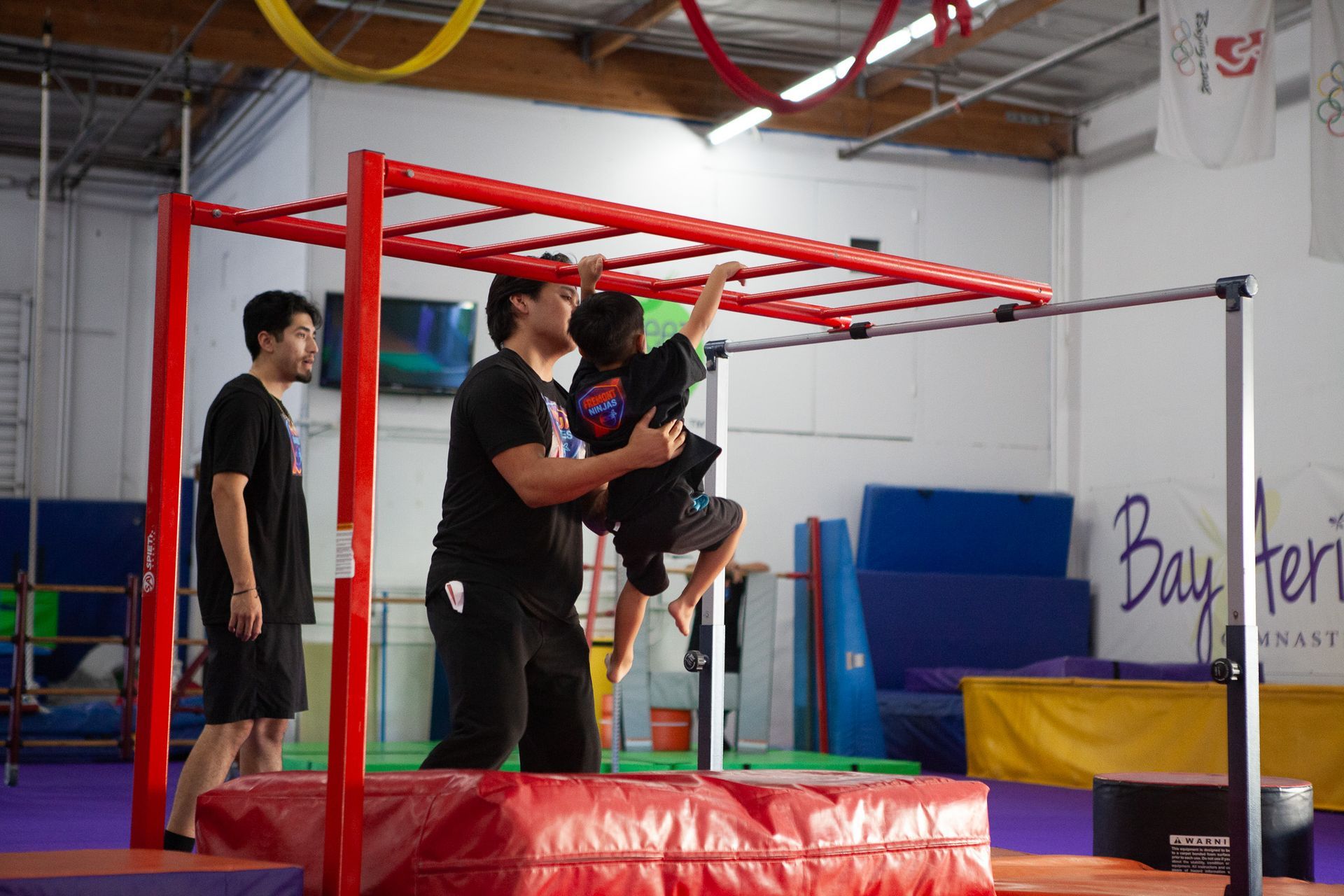 Boy hangs on red monkey bars, assisted by an adult, in a gym; another man watches.