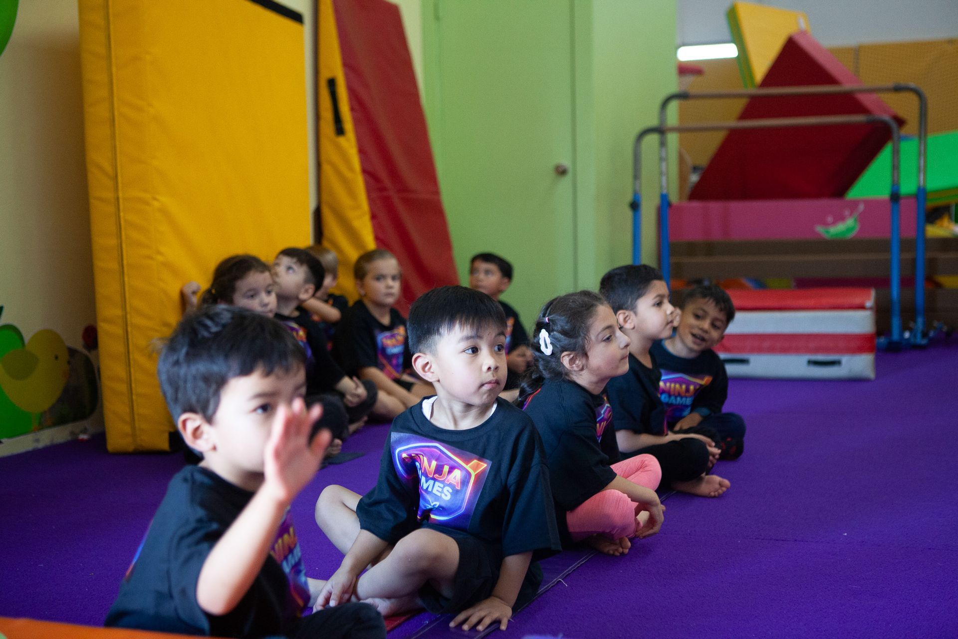 Children in black shirts sit in a row on a purple floor, some looking forward, others waving. Gym equipment in background.