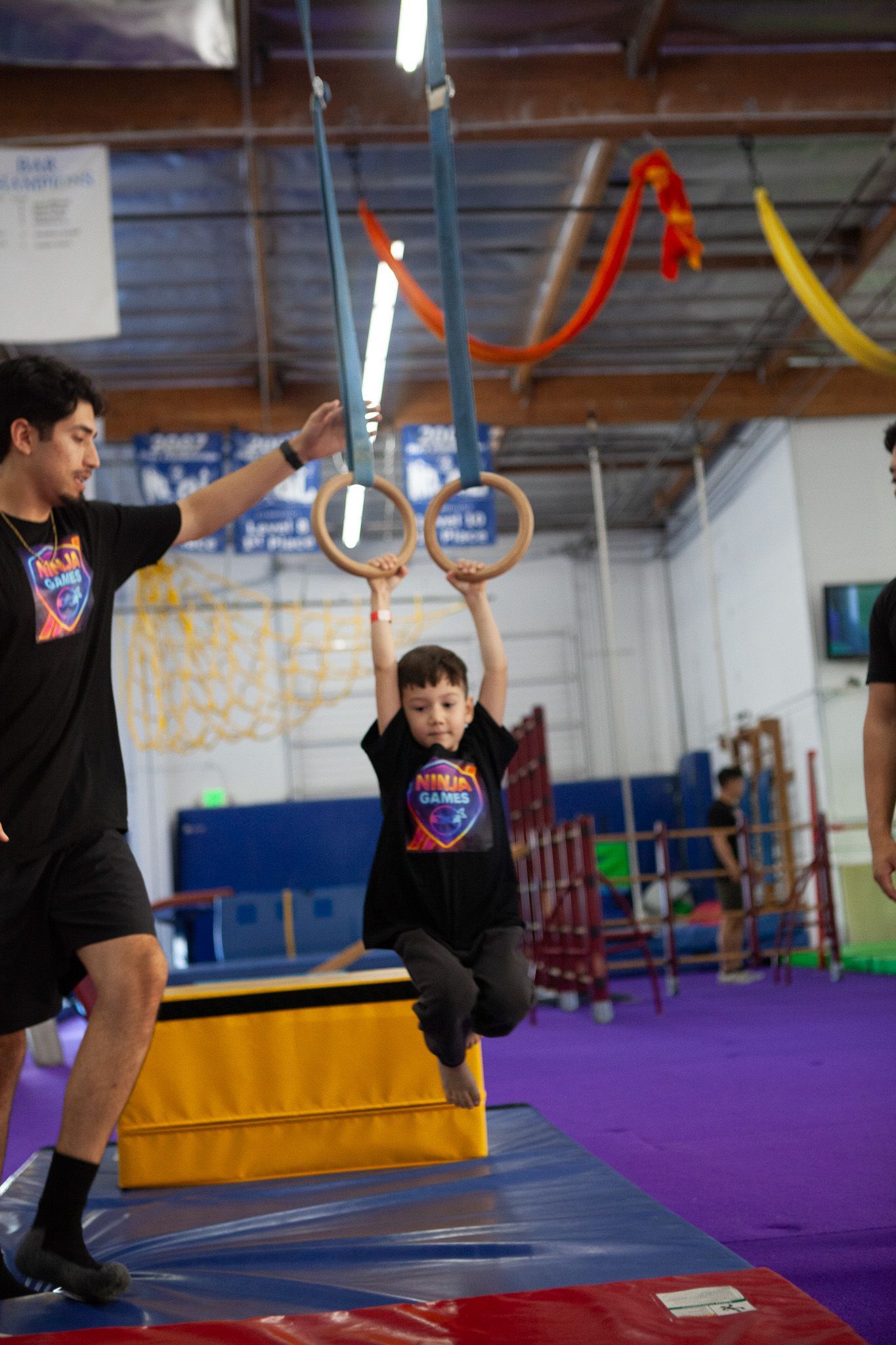 Young boy hanging from gymnastic rings, supported by an instructor in a gym setting.