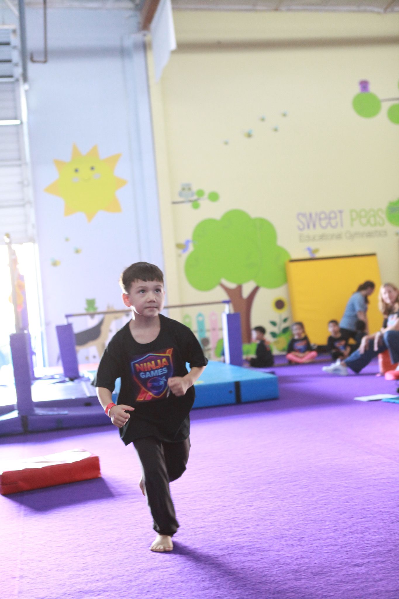 Boy running across a purple-carpeted gym floor toward the camera, other people visible in the background.
