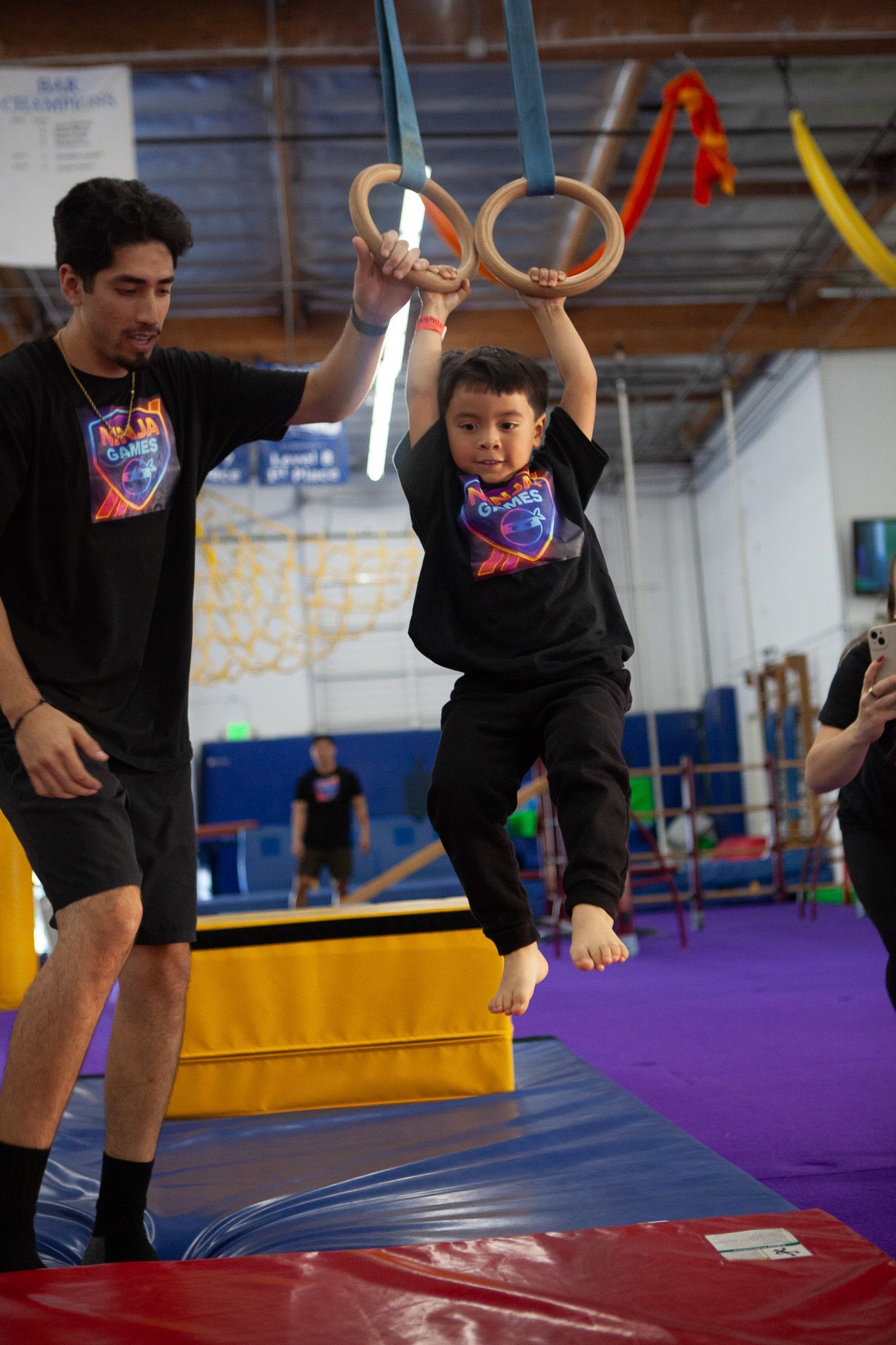Child hanging from rings with assistance from an adult in a gym.