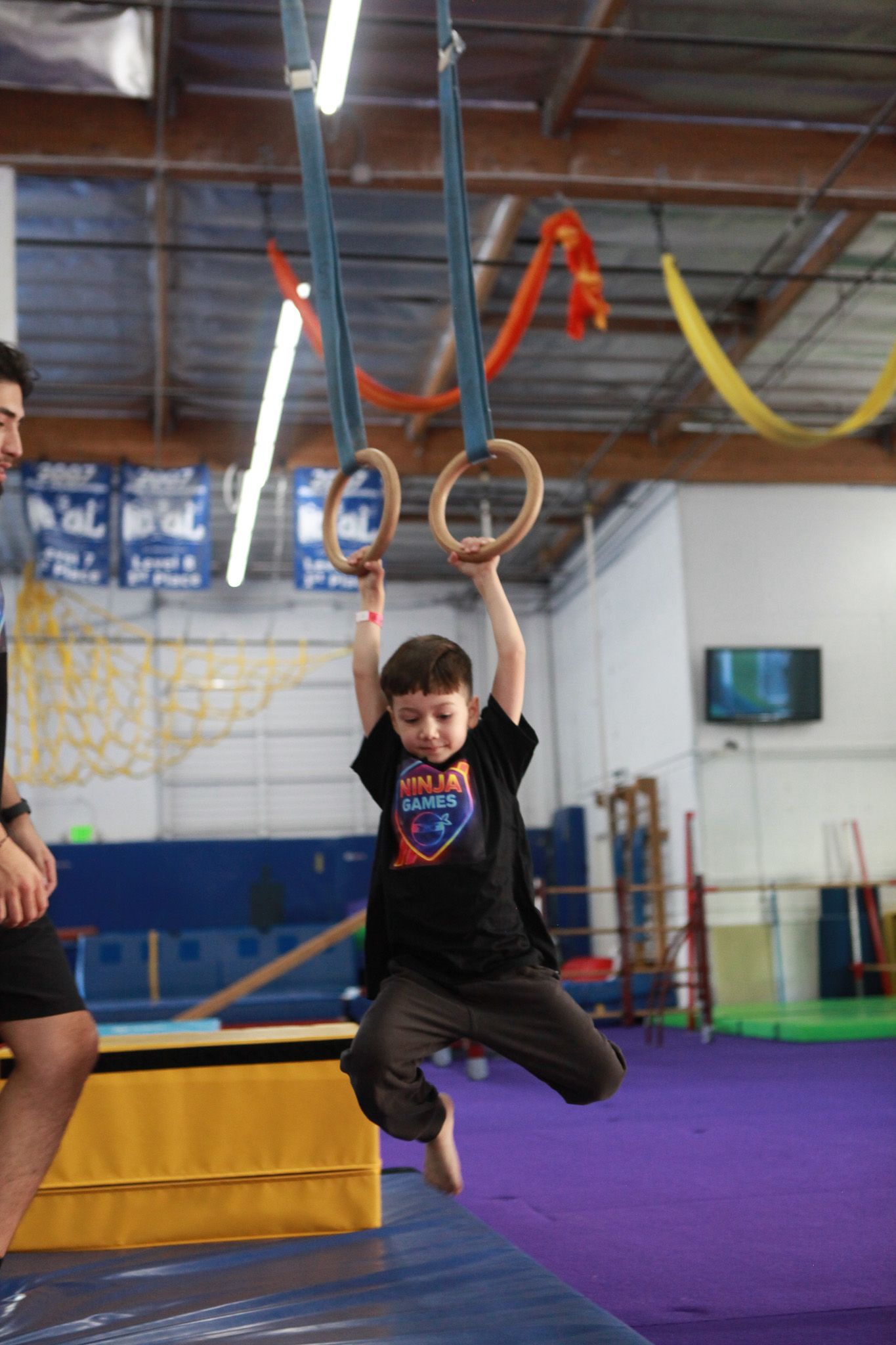 Boy hanging from gymnastic rings in a gym. A coach watches nearby.