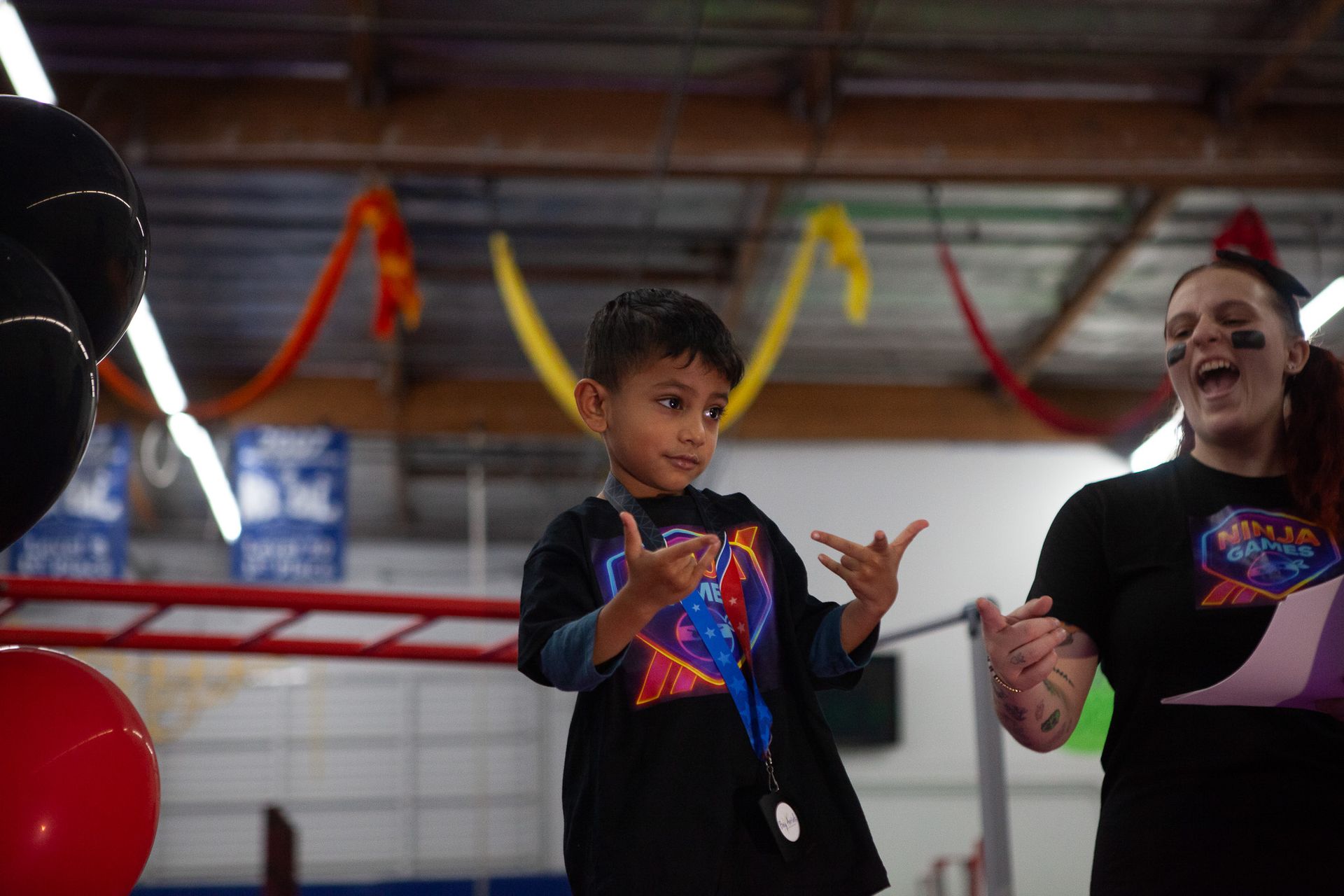Young boy with arms raised poses with a woman wearing a black t-shirt; indoor setting with balloons and ribbons.