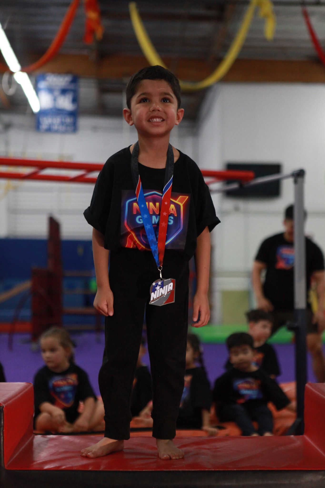 Child smiling, standing on a red mat at a Ninja Zone gym.