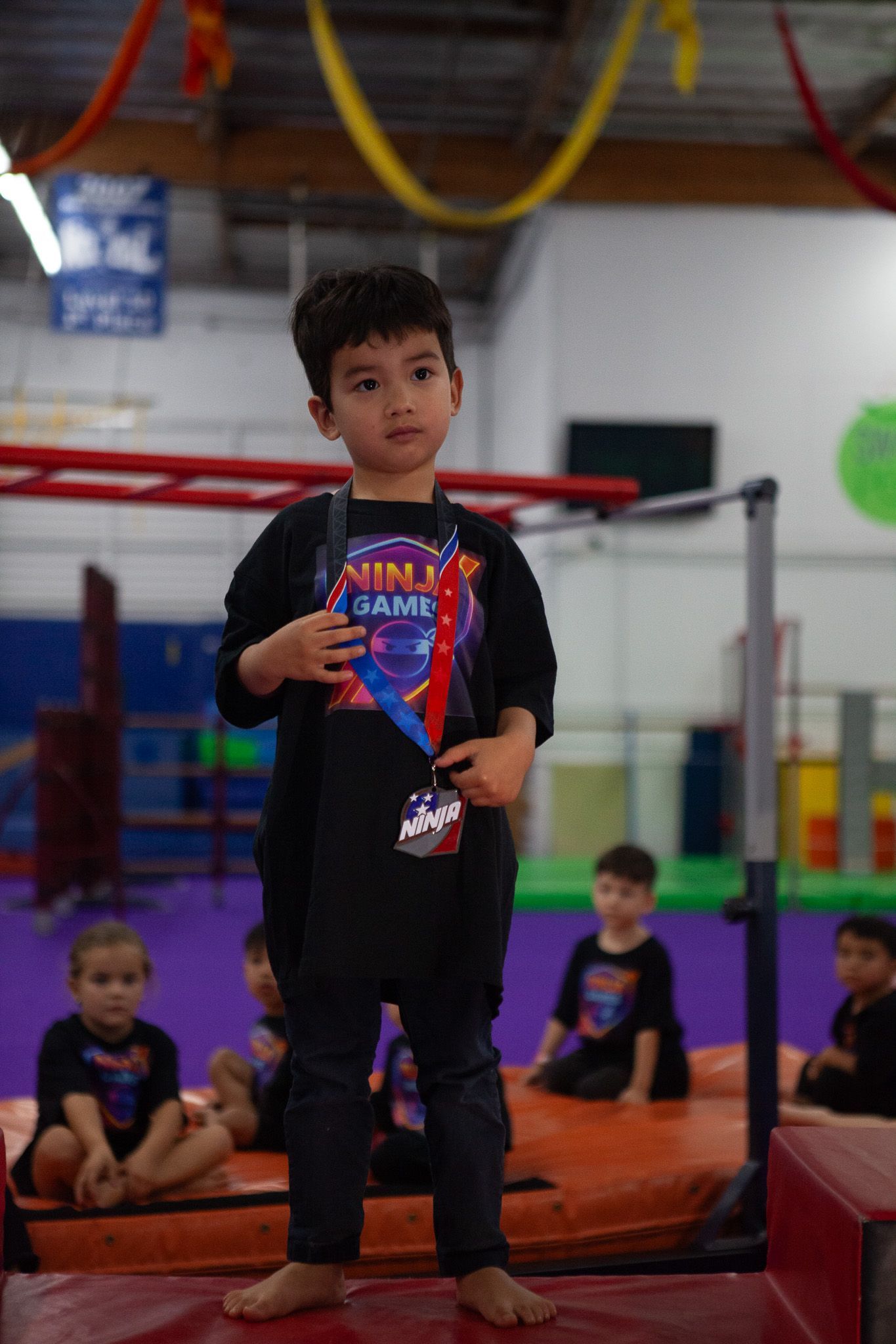 Child wearing a medal stands on a mat in a gym, several other children seated nearby.