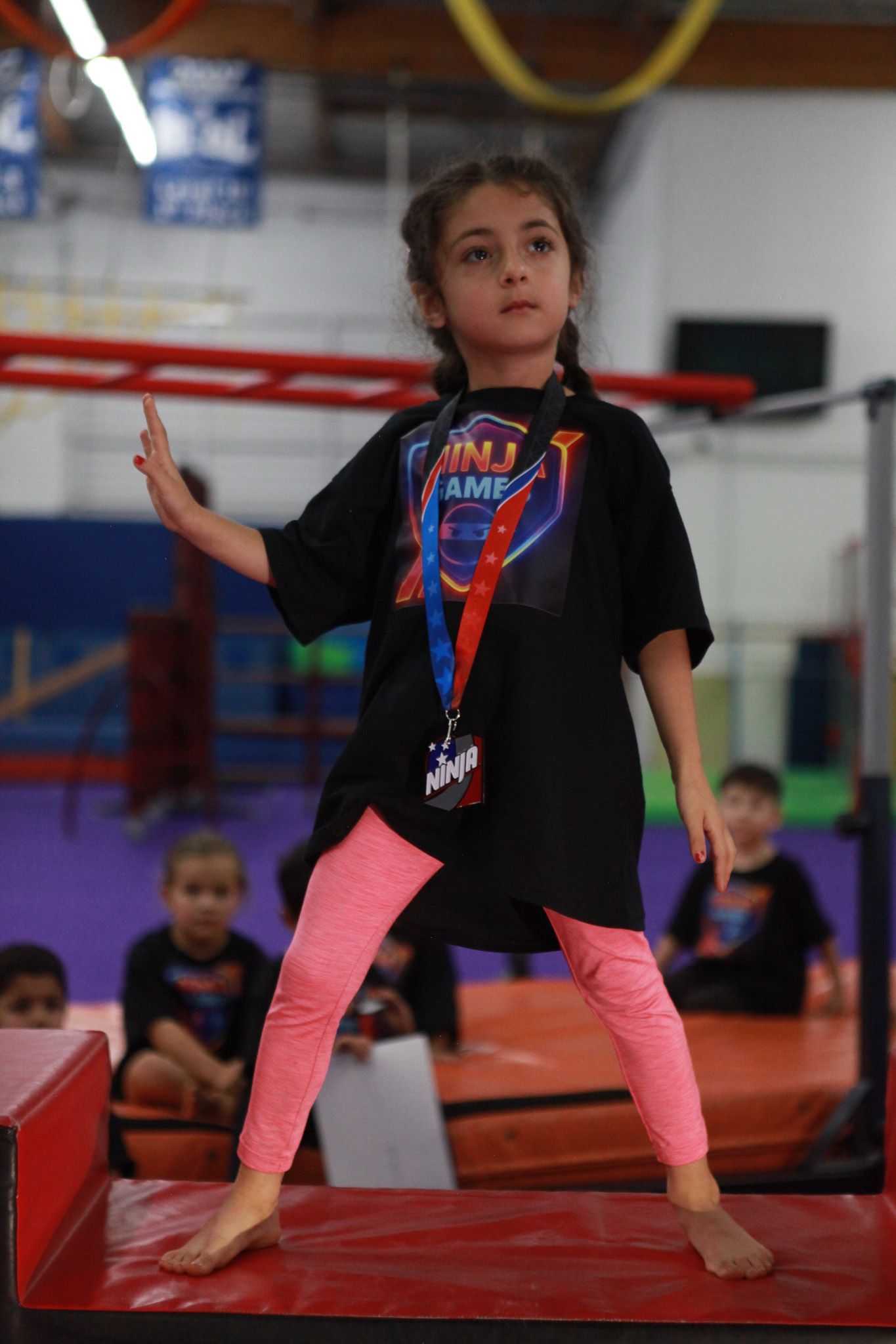 Young girl in pink leggings and black shirt stands on red platform with arm raised. Gym setting.