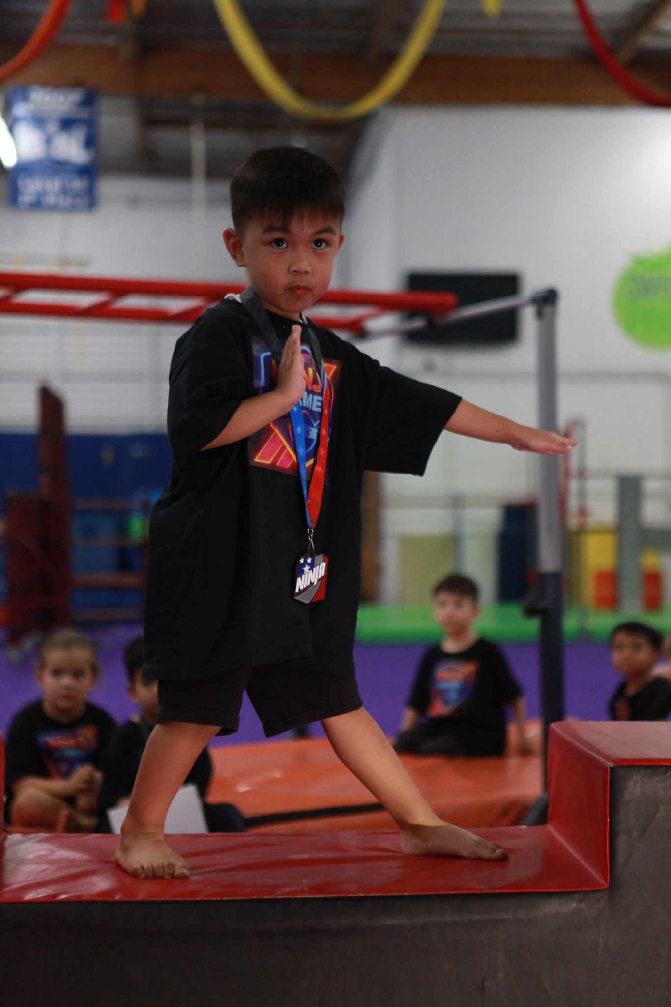 Young child in black t-shirt and shorts performs a martial arts pose on red blocks in a gym.