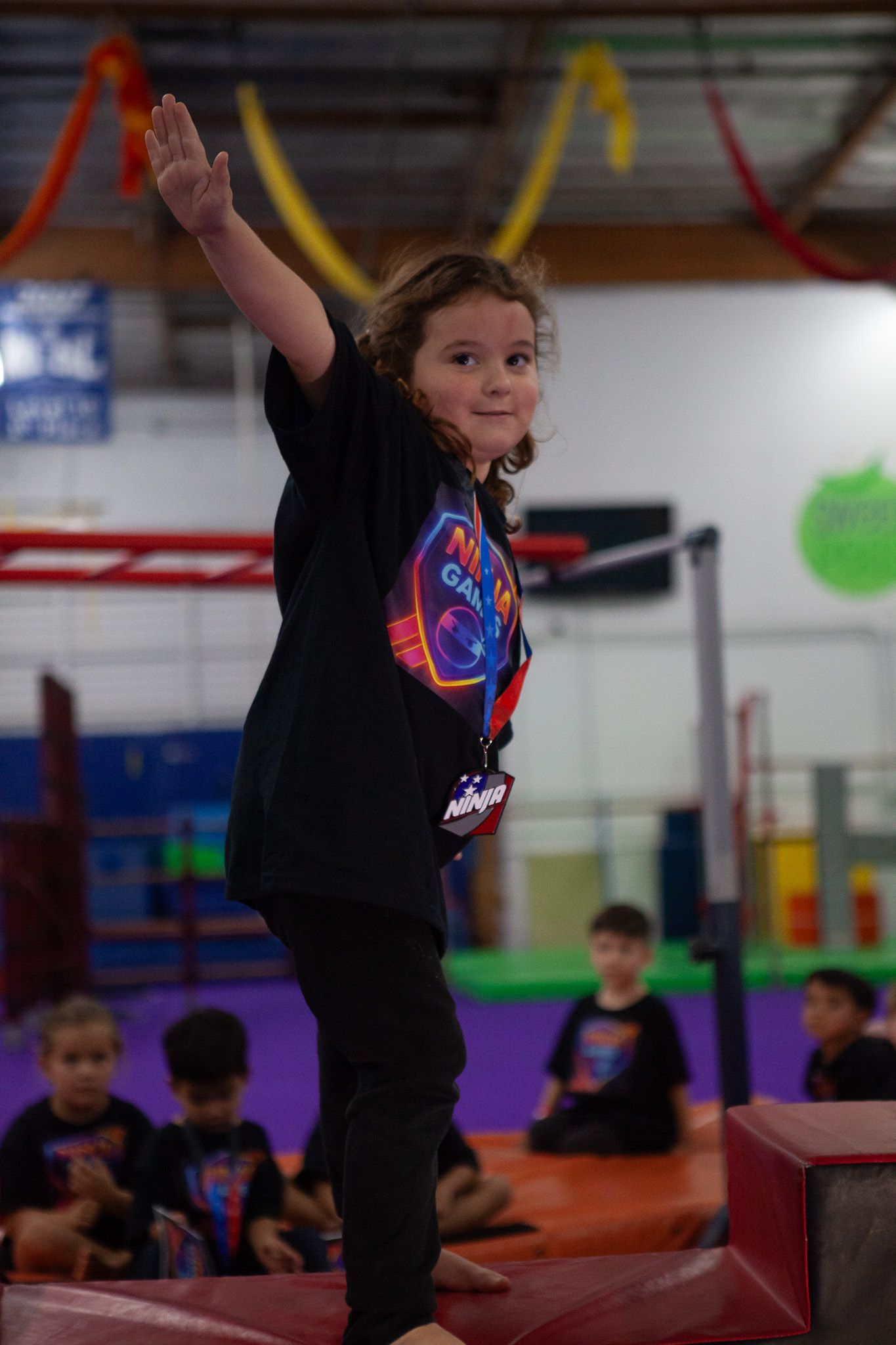 Girl balances on a beam, arm raised, smiles. Children watch in a gym.