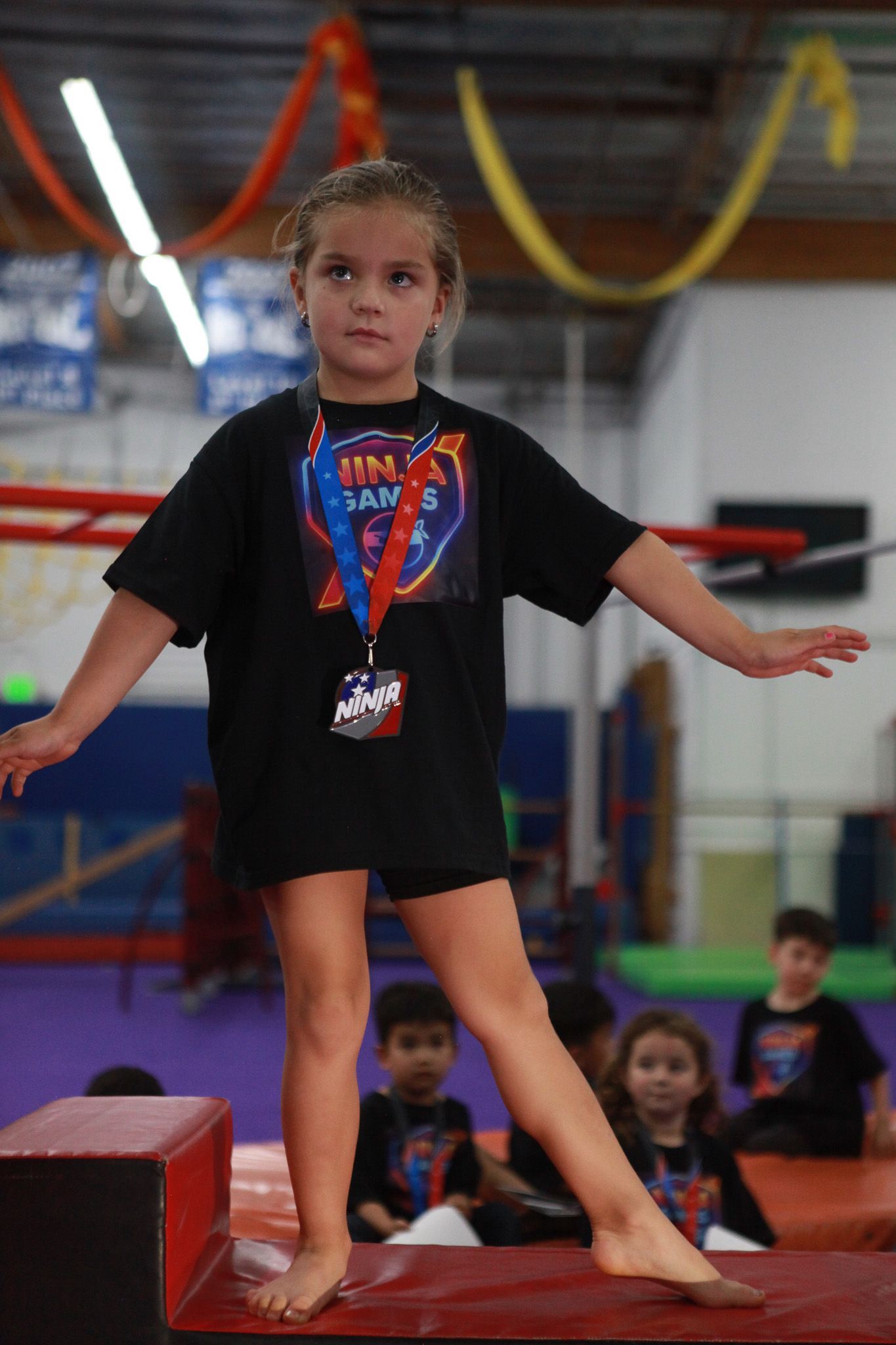 Young girl balancing on a beam in a gymnasium, wearing a black shirt and medal.