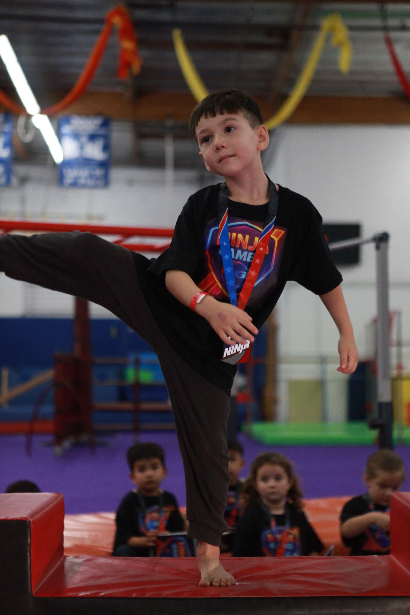 Boy in a Superman shirt doing a martial arts kick in a gym, other kids watch.