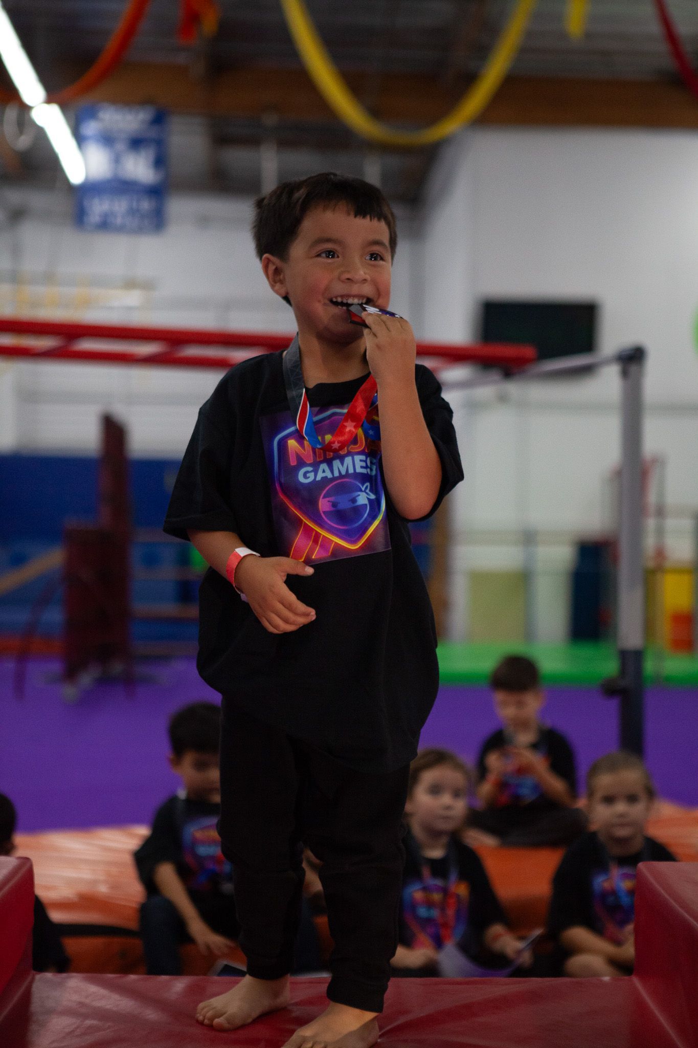 Boy with medal standing on mat; others seated. Gym with colorful decorations.