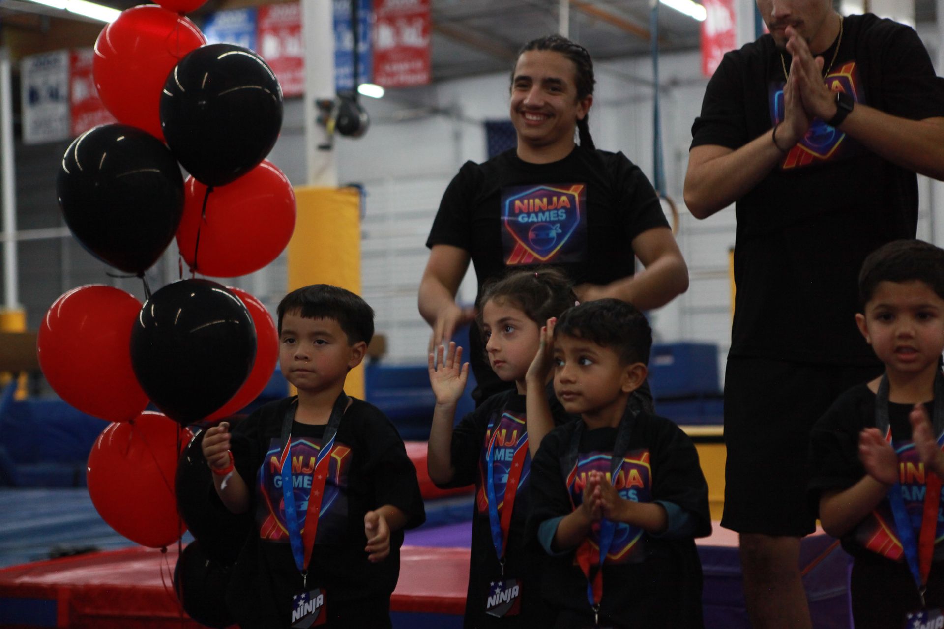 Children in black shirts with medals, holding balloons, celebrate in a gym. Adults clap behind them.