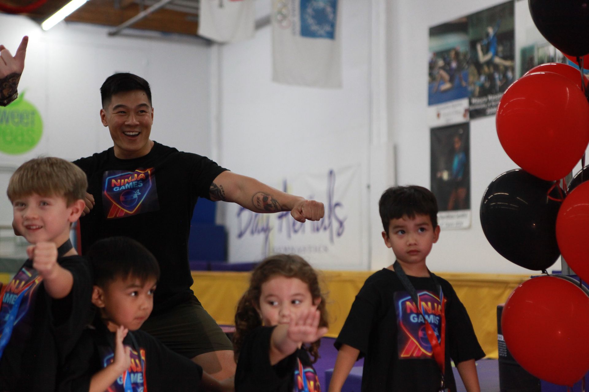 Man with children in a gym, all wearing black shirts. Some are giving fist bumps. Red and black balloons are present.