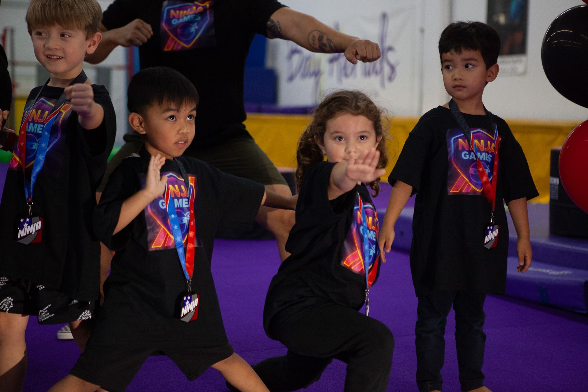Children in black shirts with medals pose martial arts stances on a purple mat.
