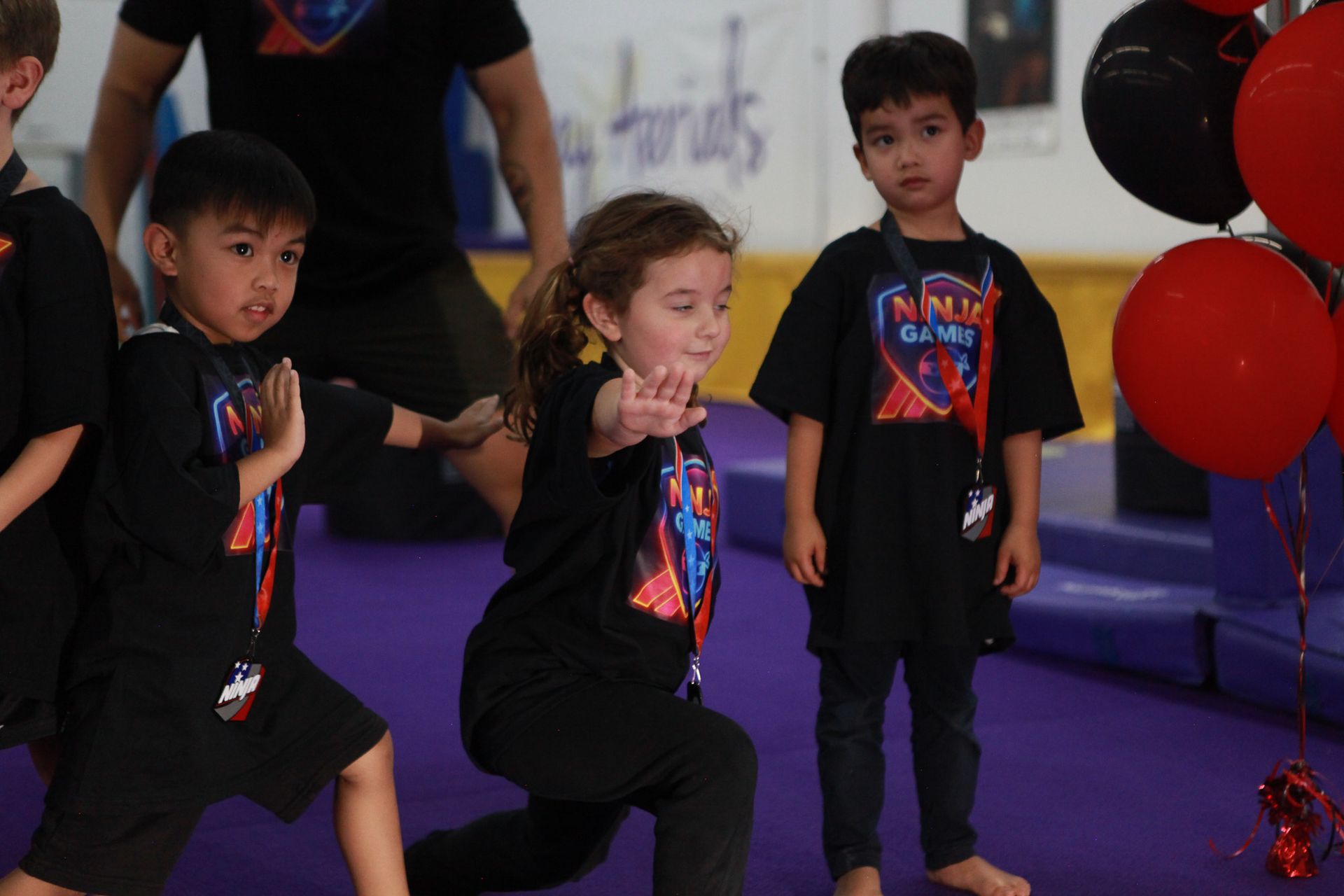 Children in black t-shirts practicing martial arts on a purple mat, red and black balloons in the background.