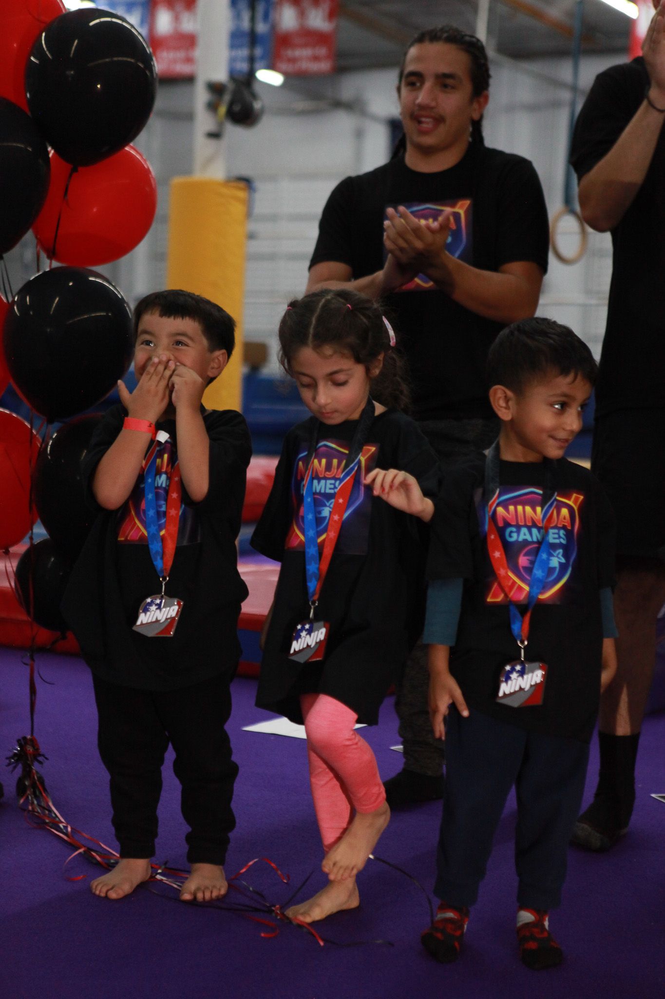 Children at a gymnastics event stand, celebrating. Balloons and adults in the background.