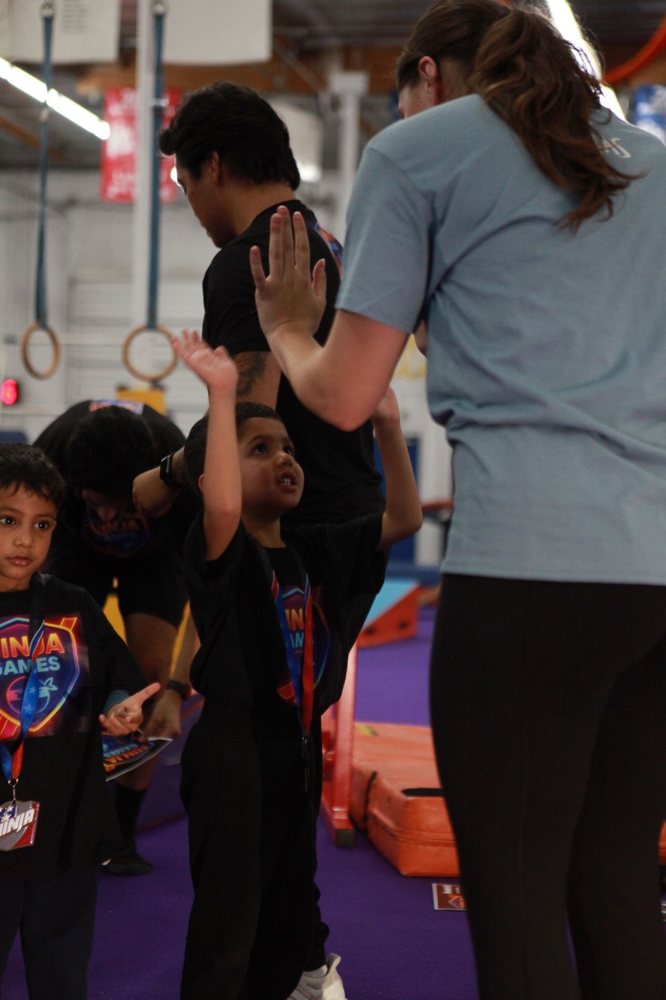 Boy raising hand at a gymnastics class, teacher high-fiving him. Other kids in background.