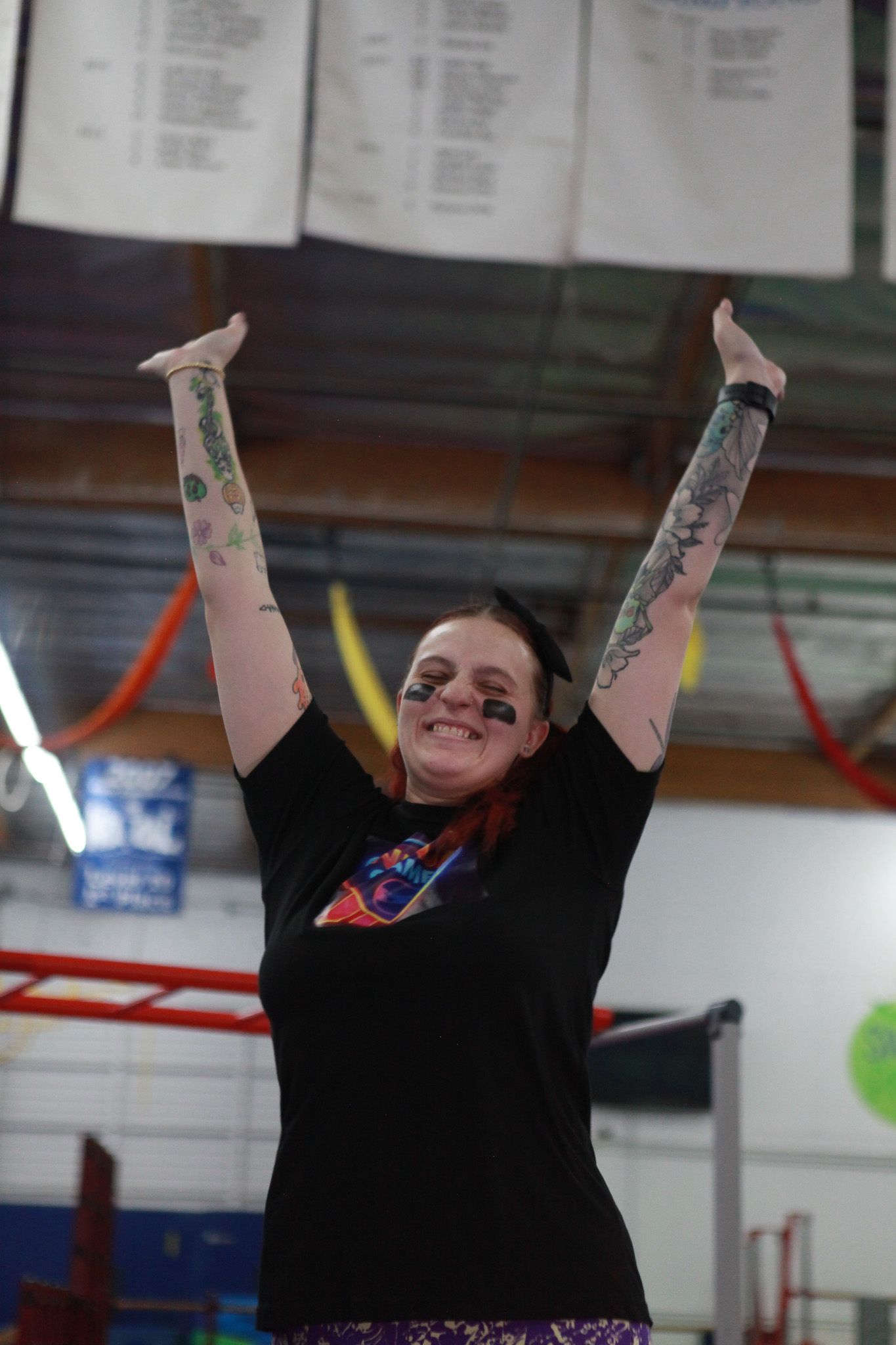 Person with tattoos raising arms in celebration at an indoor sports venue. Black t-shirt, eye black.