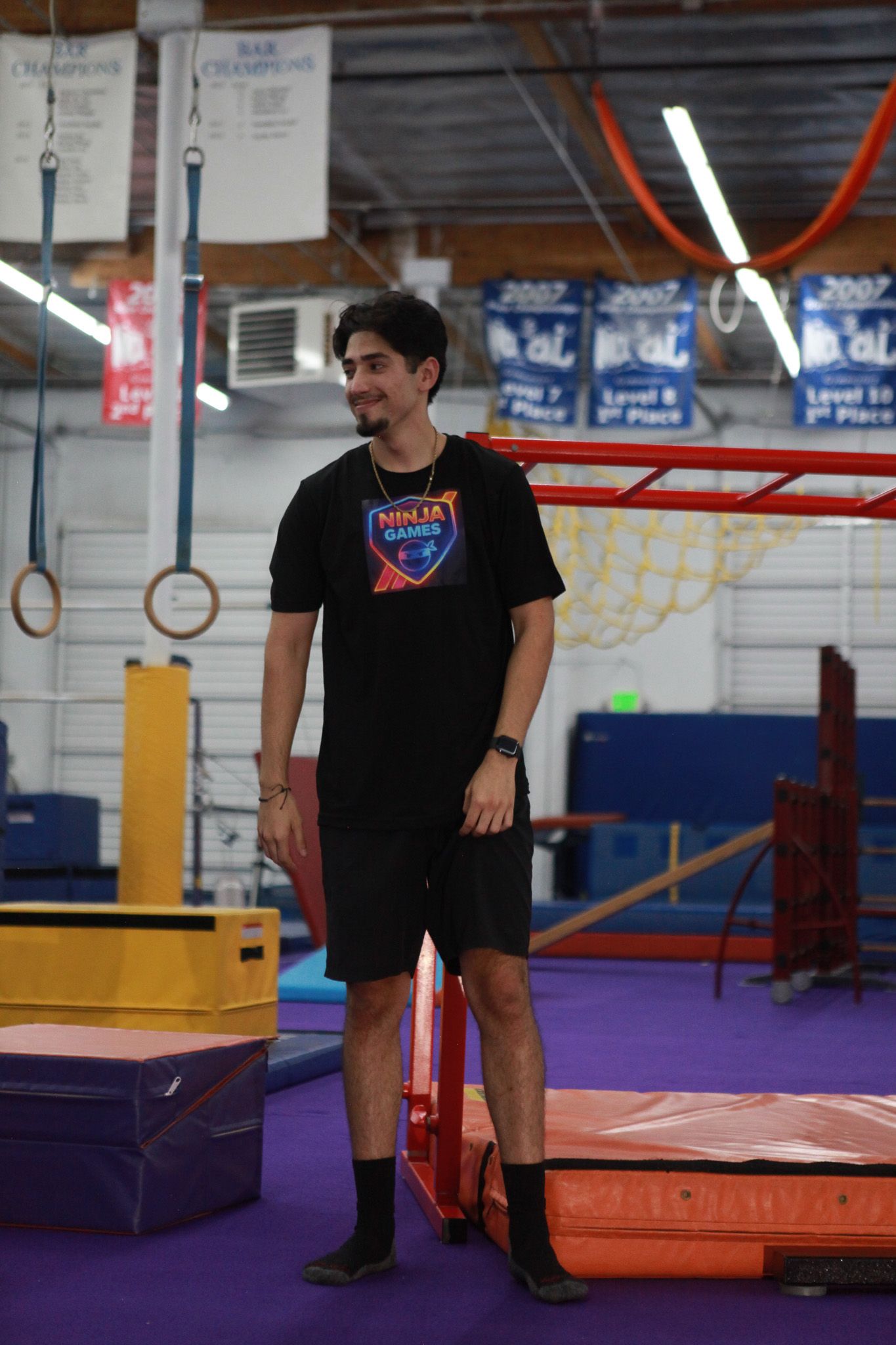 Man in black clothing smiles in a gym, near obstacle course equipment.