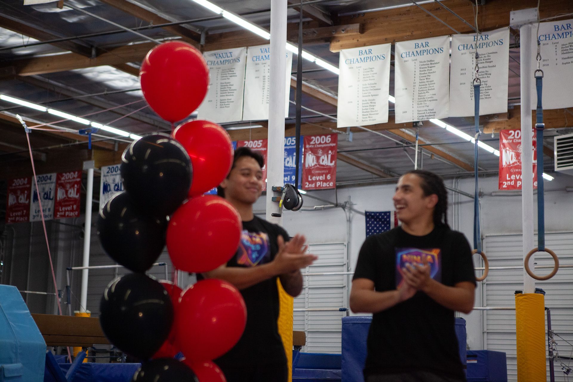 Two men clapping next to a balloon arrangement in a gym. Black and red balloons.