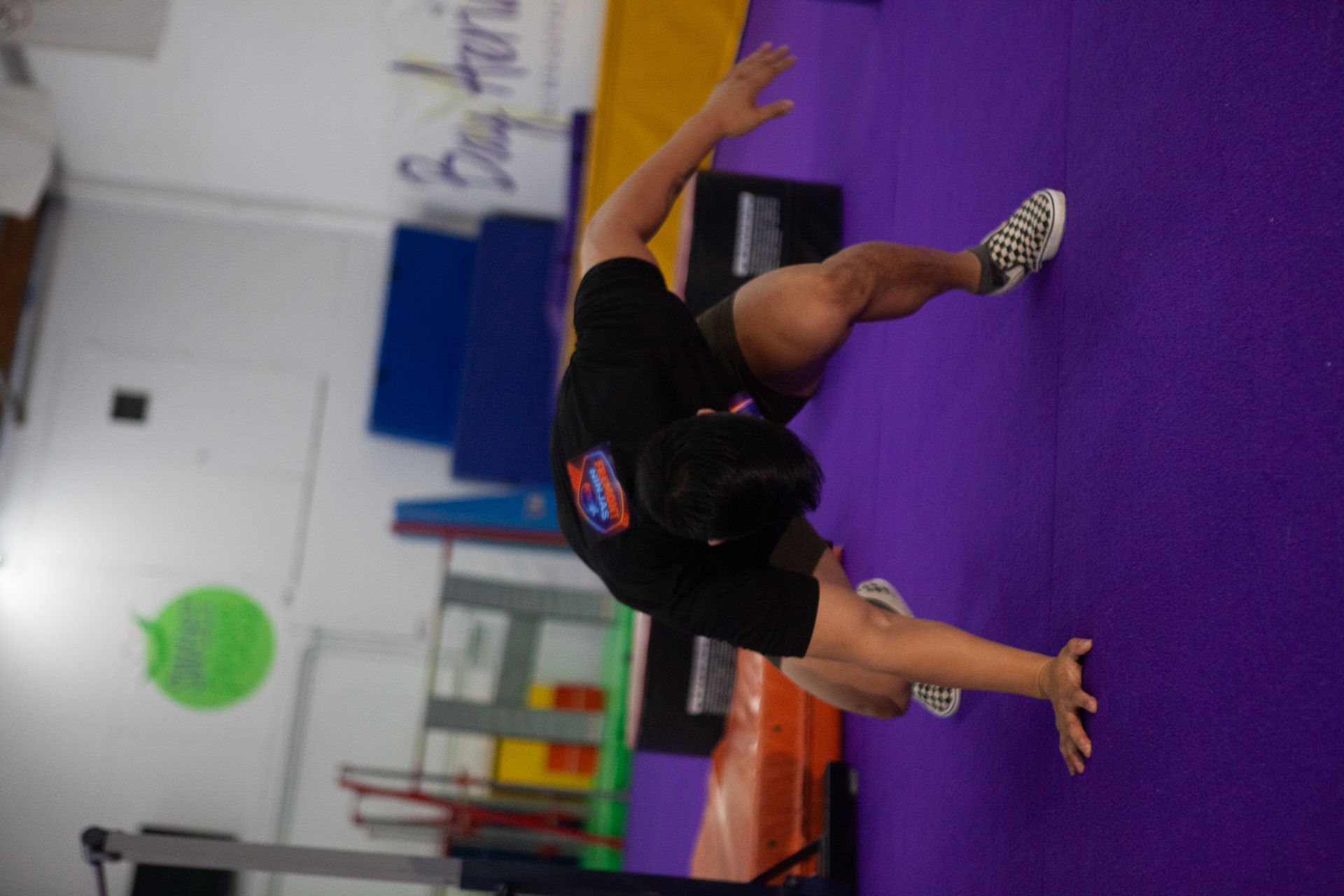 Person in black shirt, stretching on purple mat, arms outstretched. Gym setting.