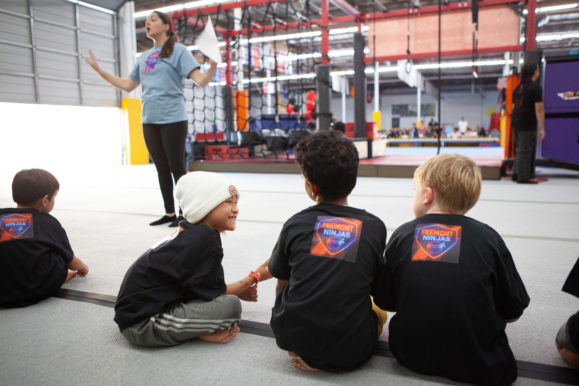 Children in black shirts listen to an instructor in a gym setting.