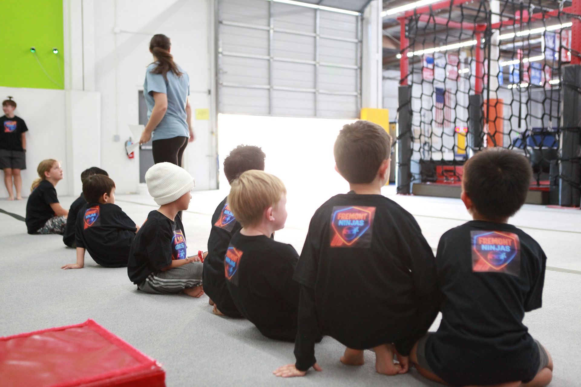 Children in black shirts, kneeling and watching a woman in a gym. Red block in front.