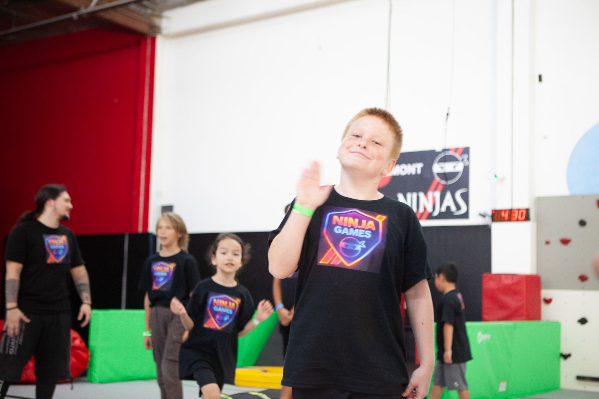 Boy in black shirt waves in a ninja gym. Others in matching shirts and an instructor are visible.
