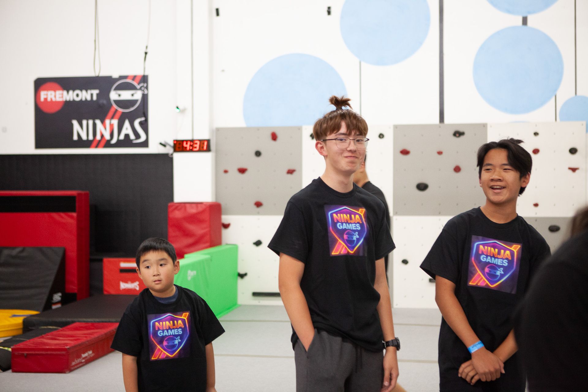 Three young people wearing matching black shirts stand in a gym, with a wall of climbing holds.