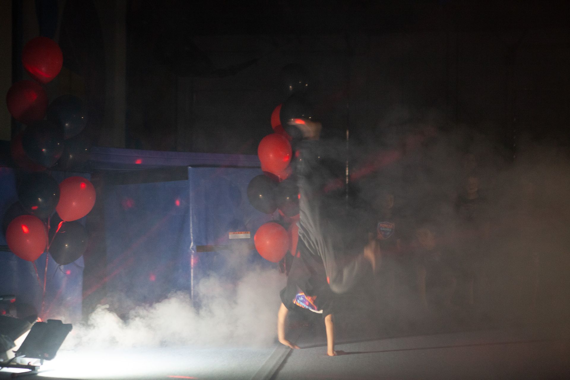 A person performing a handstand on a stage, with balloons and fog in the background.