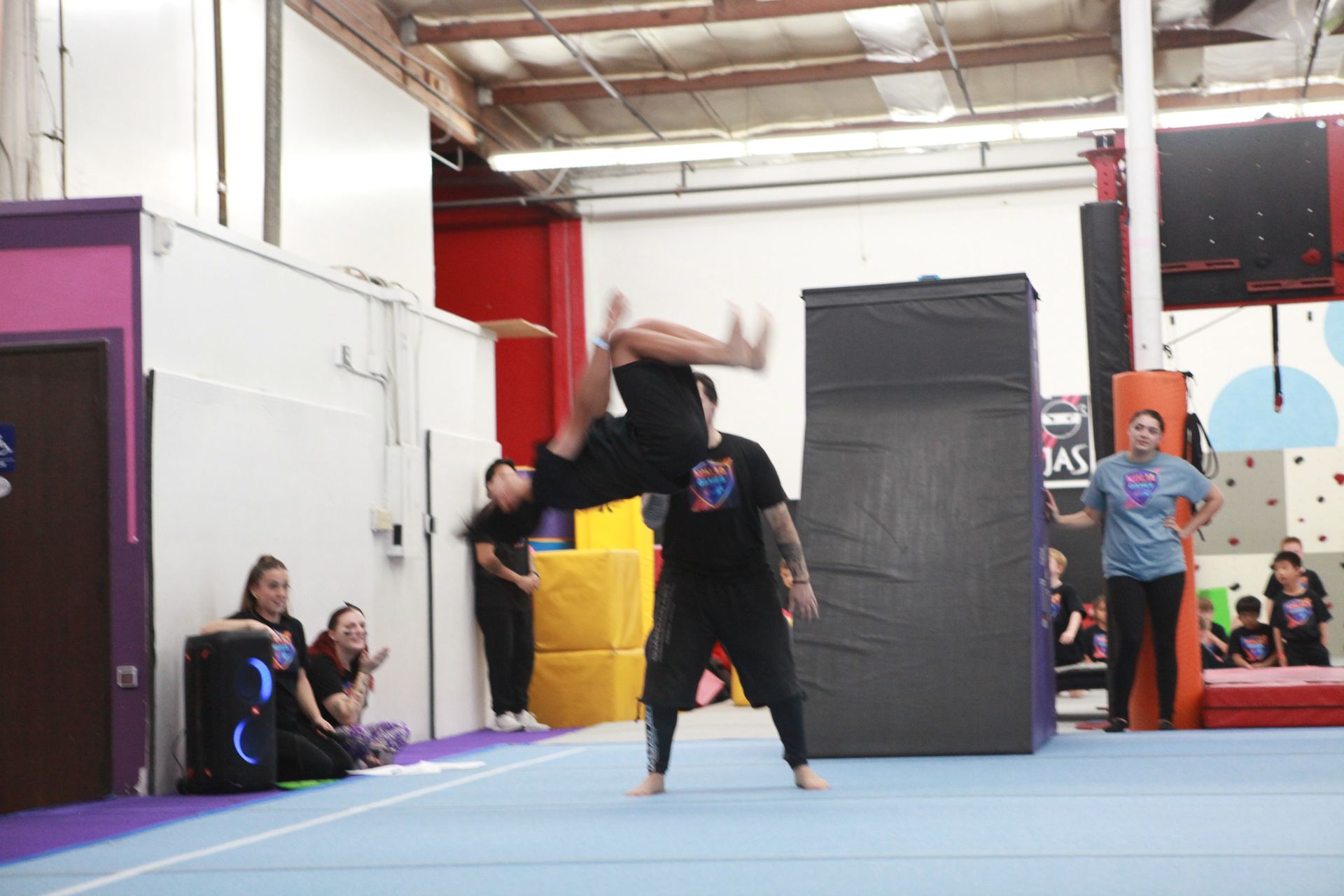 A person performing a backflip, assisted by another, inside a gym. Audience watches.