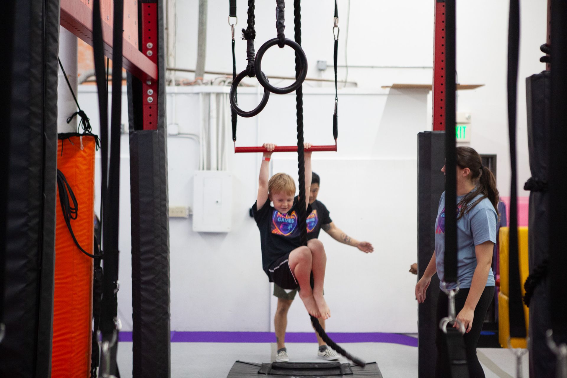 Boy climbing a rope at an indoor obstacle course, assisted by two adults.
