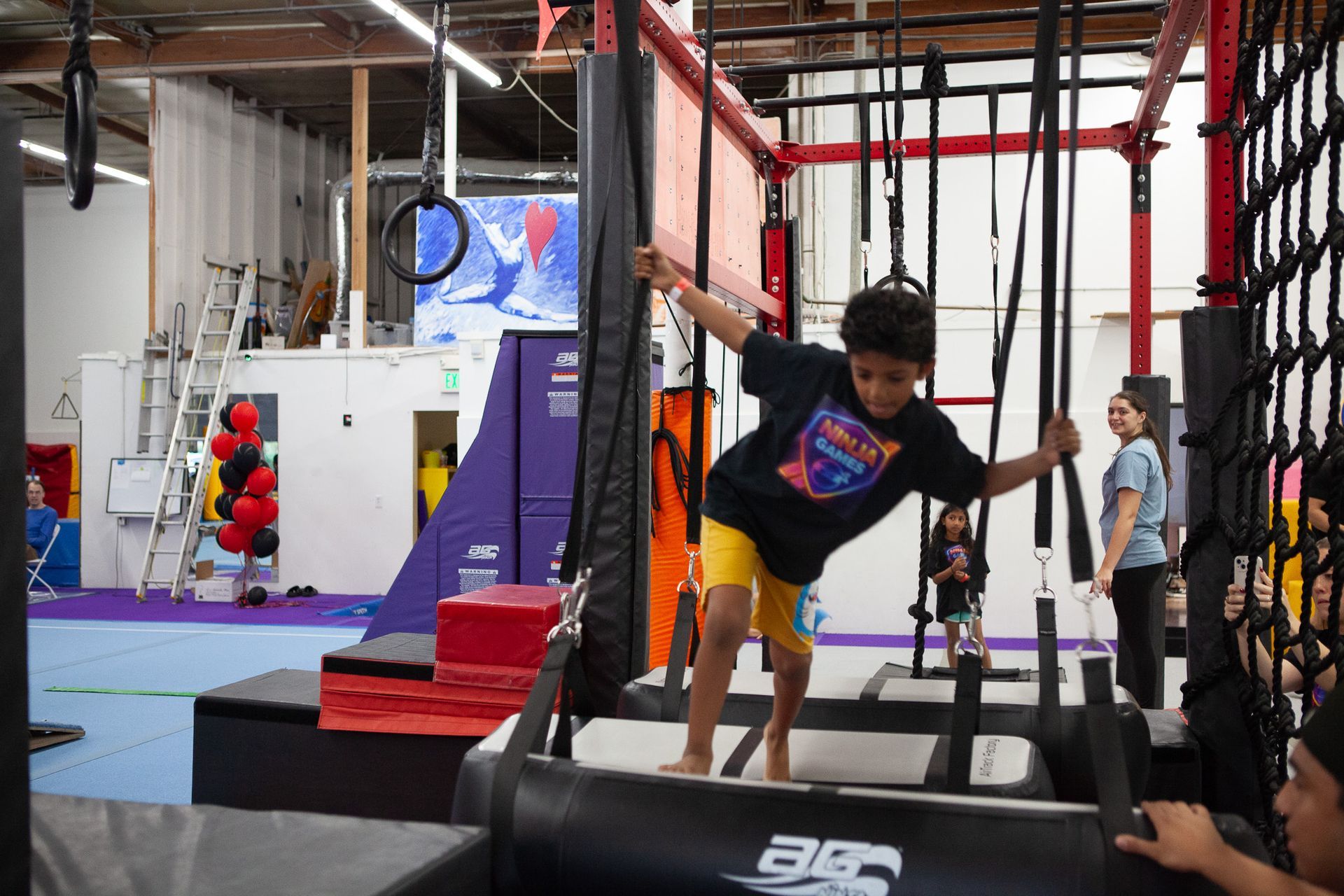 Boy navigating an obstacle course, holding onto bars, yellow shorts, indoor gym setting.