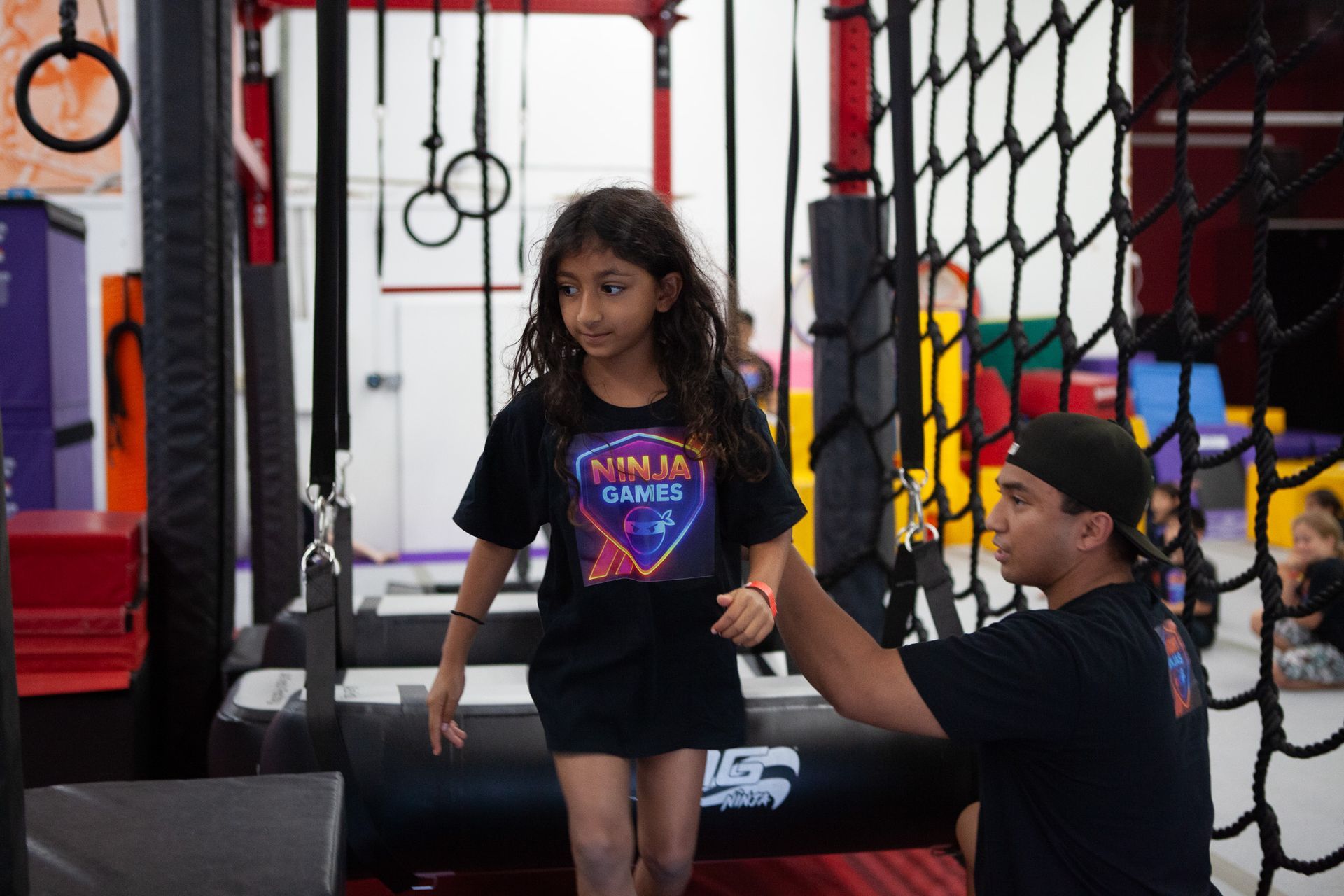 Girl in black shirt at a ninja gym, assisted by a coach. She's navigating an obstacle course.