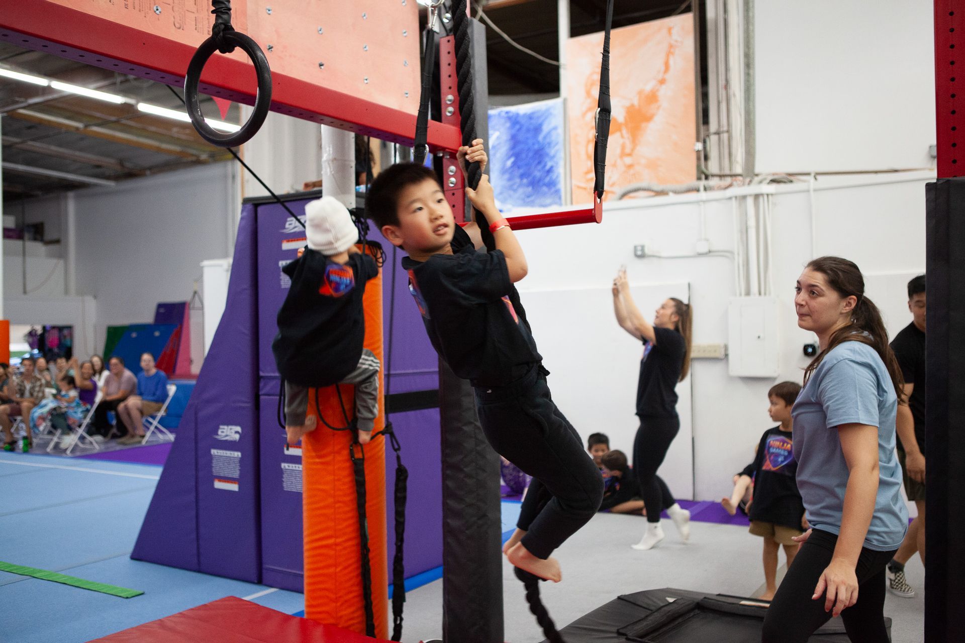 Boy hanging from a rope at an obstacle course, another child climbing, adults and children observing.