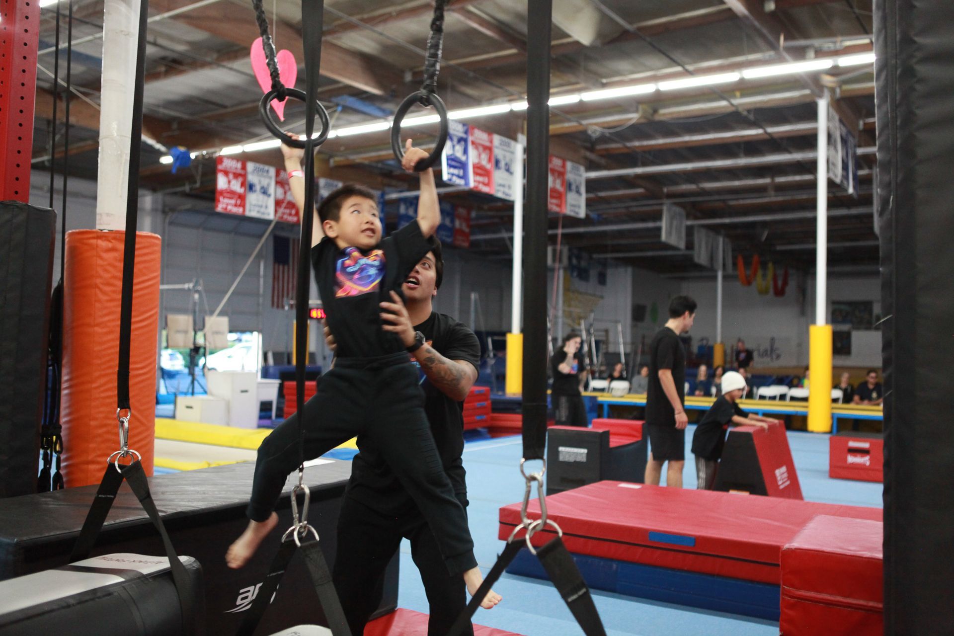 Boy hanging from rings, assisted by an adult, in a gym.