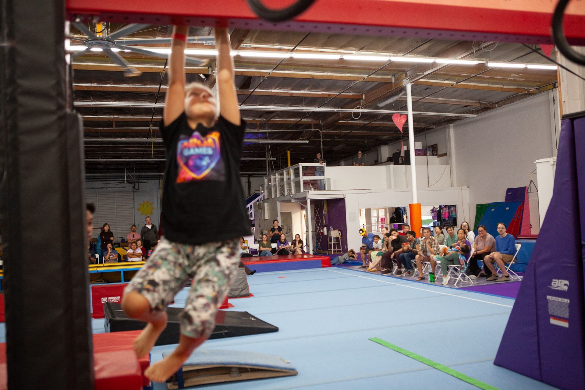 Person hanging from a red overhead bar in a gym setting, holding on with both hands.