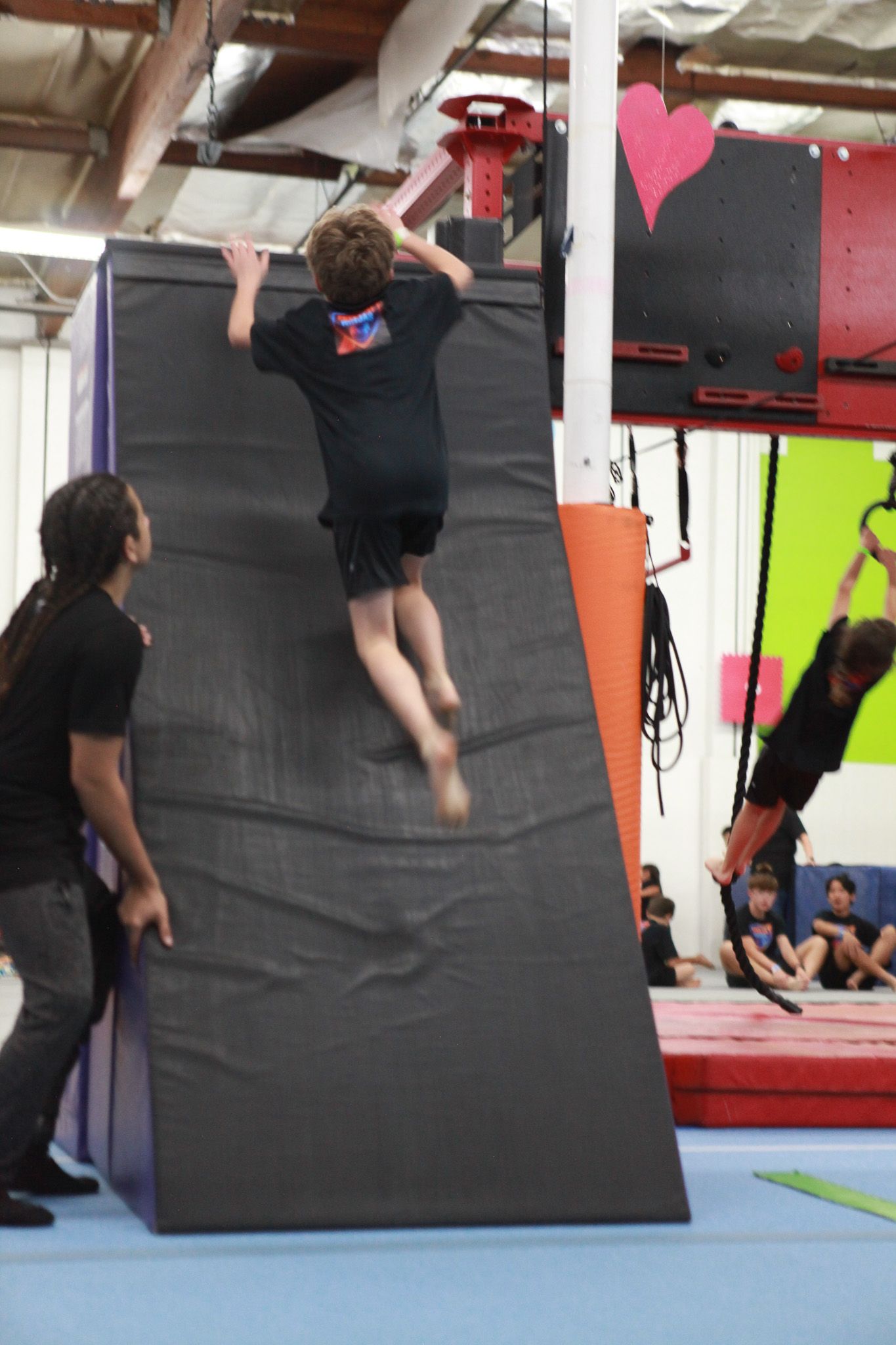 Boy jumps up a slanted padded wall with a coach assisting at an obstacle course gym.