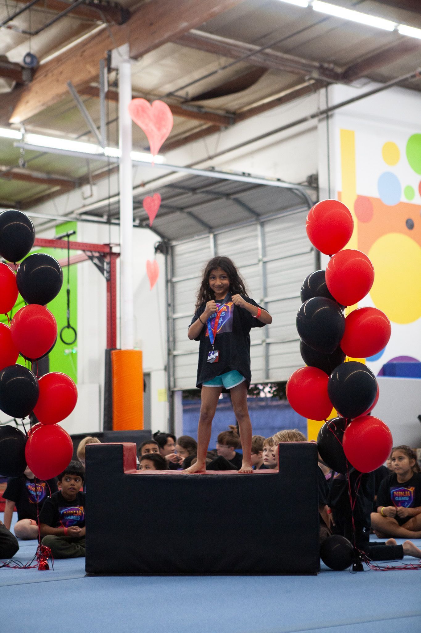 Young girl on a podium, fists up, surrounded by red and black balloons, inside a gym.